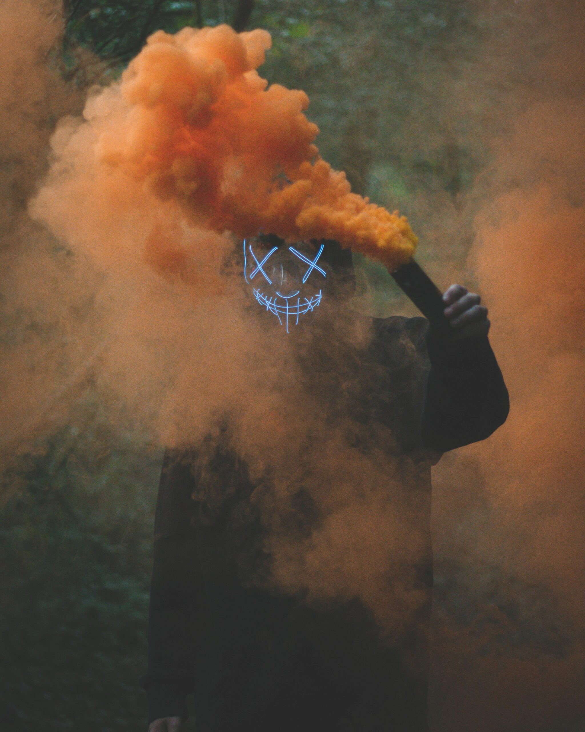 Formación A person wearing an LED mask holds a smoke bomb, enveloped in vivid orange smoke.